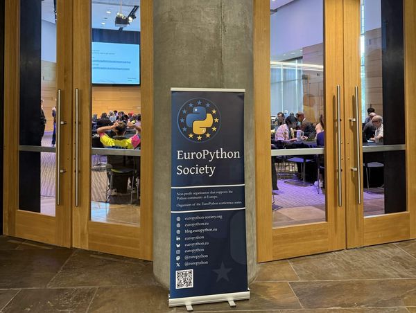 A photo of a EuroPython Society banner in an event venue, with wooden glass doors through which attendees can be seen